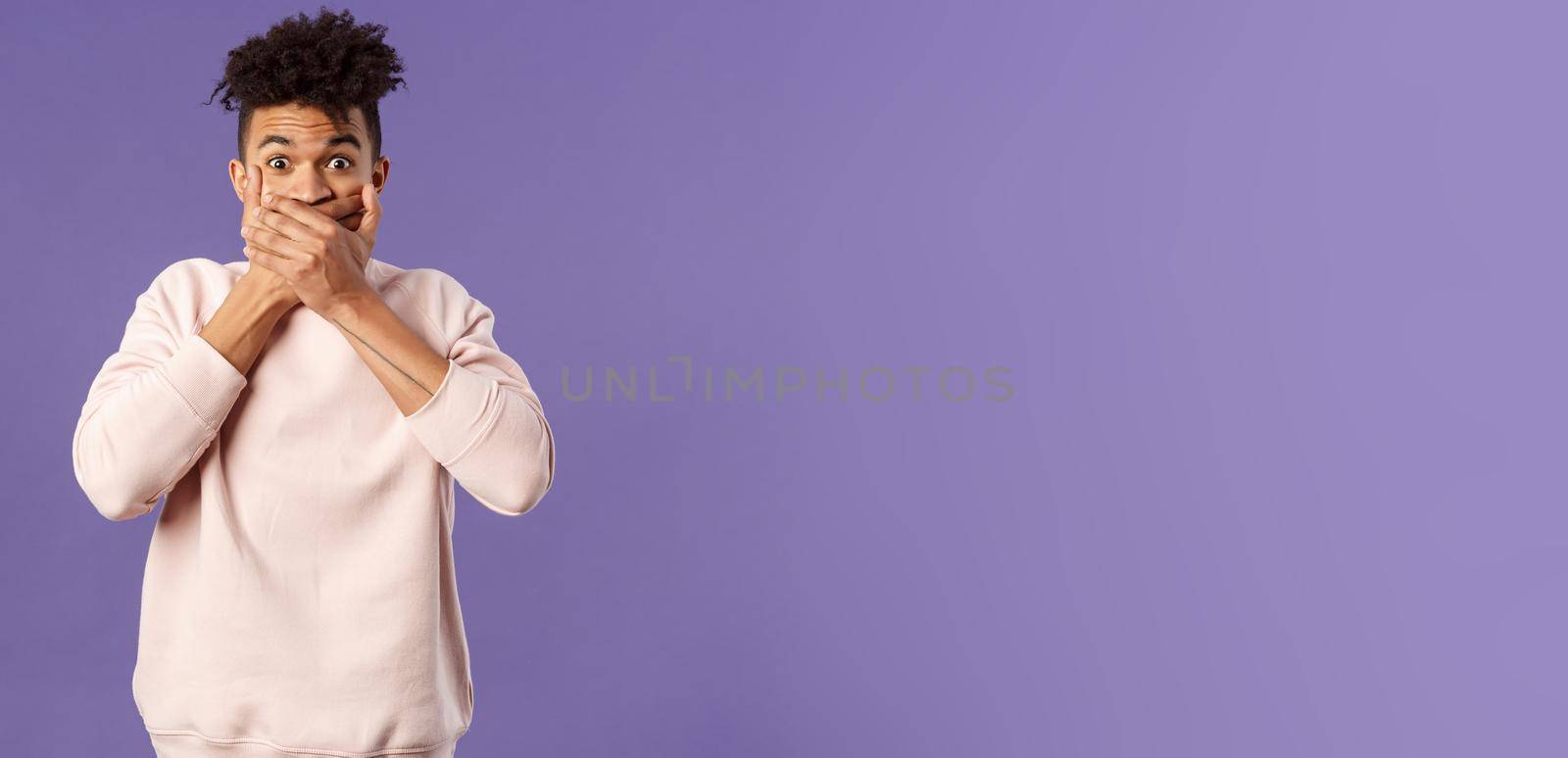 Portrait of shocked impressed young hispanic man found out gossip, heard rumour, trying not laugh shut his mouth with hands and express amazement and surprised with gaze, purple background.