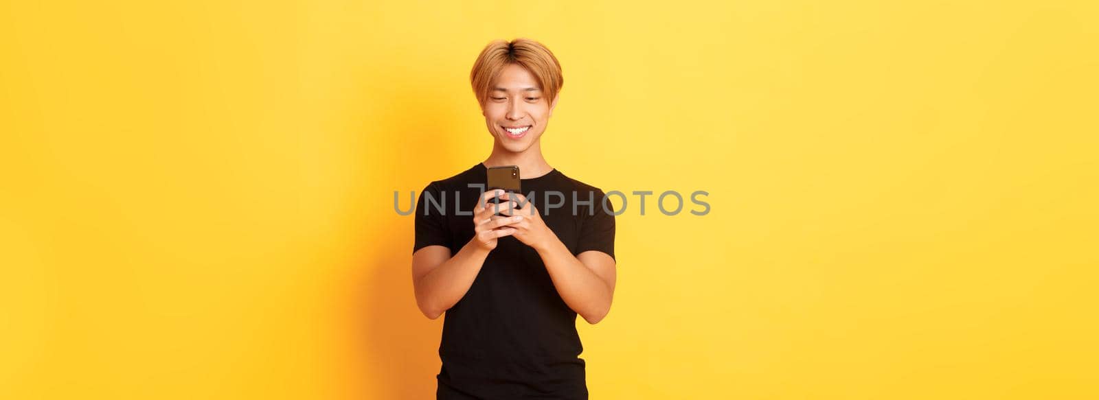 Portrait of handsome stylish asian guy with blond hair, using mobile phone and smiling, yellow background.