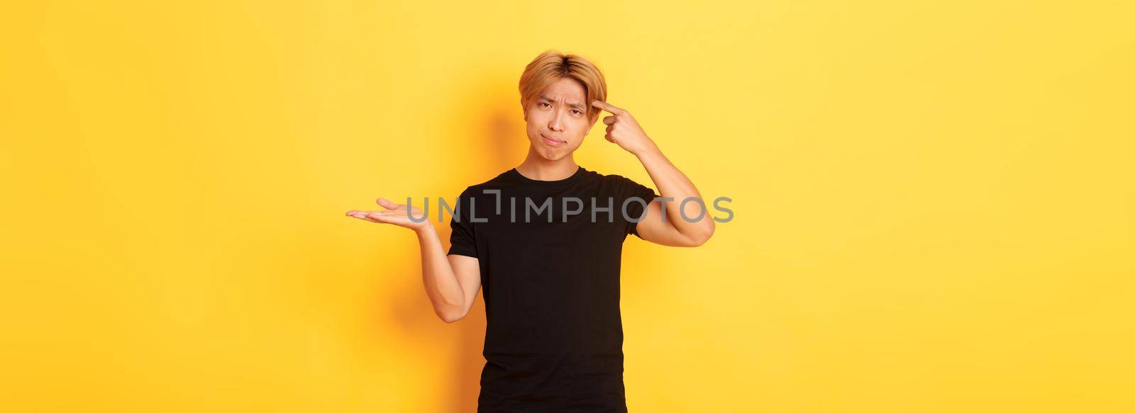 Disappointed asian guy with blond hair, raising hand up puzzled and scolding someone acting stupid, standing yellow background.