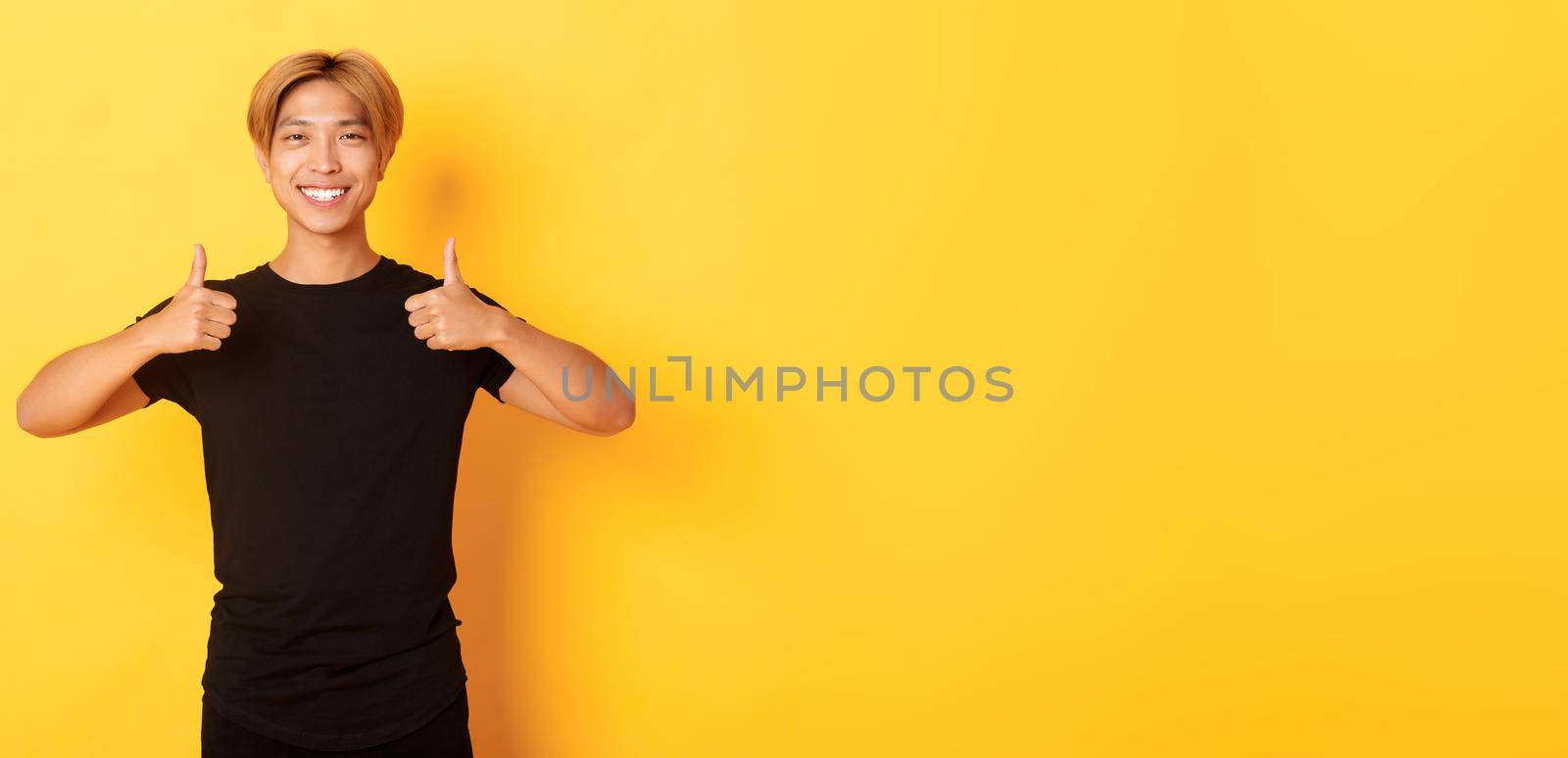 Portrait of satisfied handsome asian male student showing thumbs-up in approval, standing yellow background.