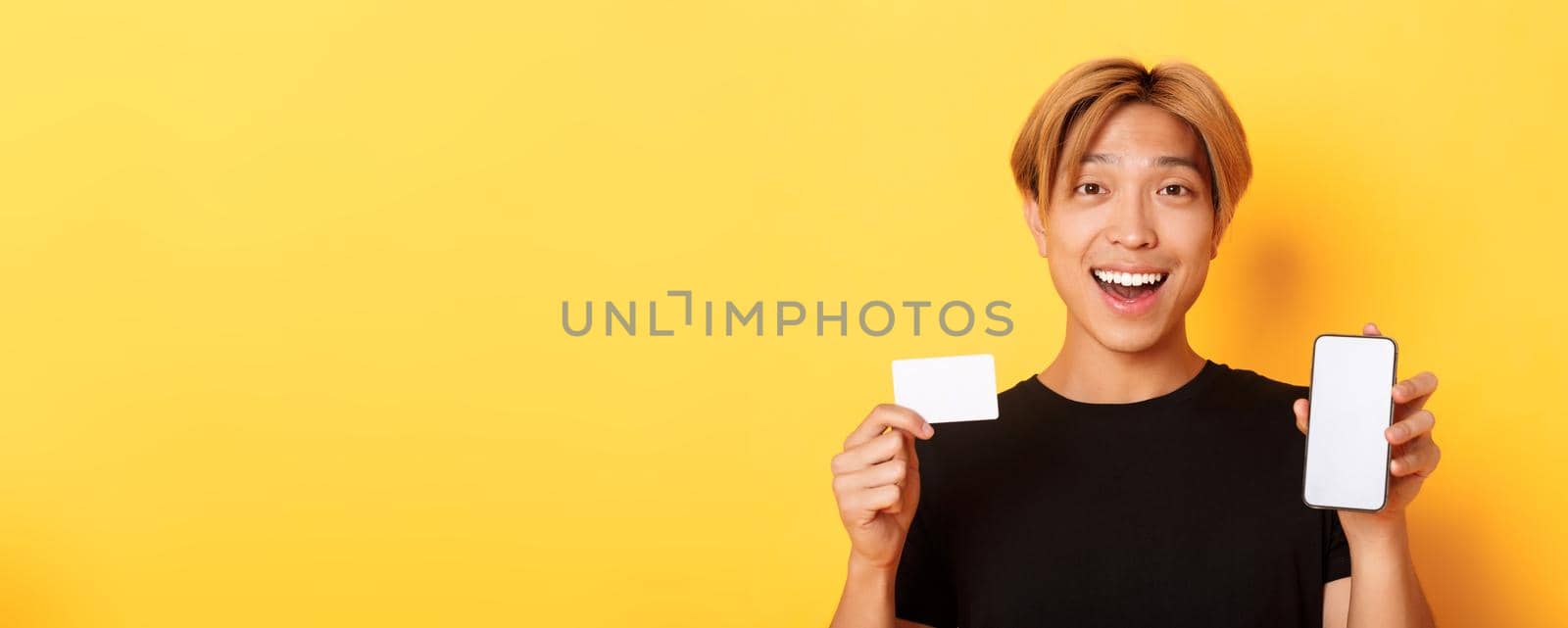 Amazed happy asian guy showing credit card and smartphone screen, smiling fascinated, standing yellow background.