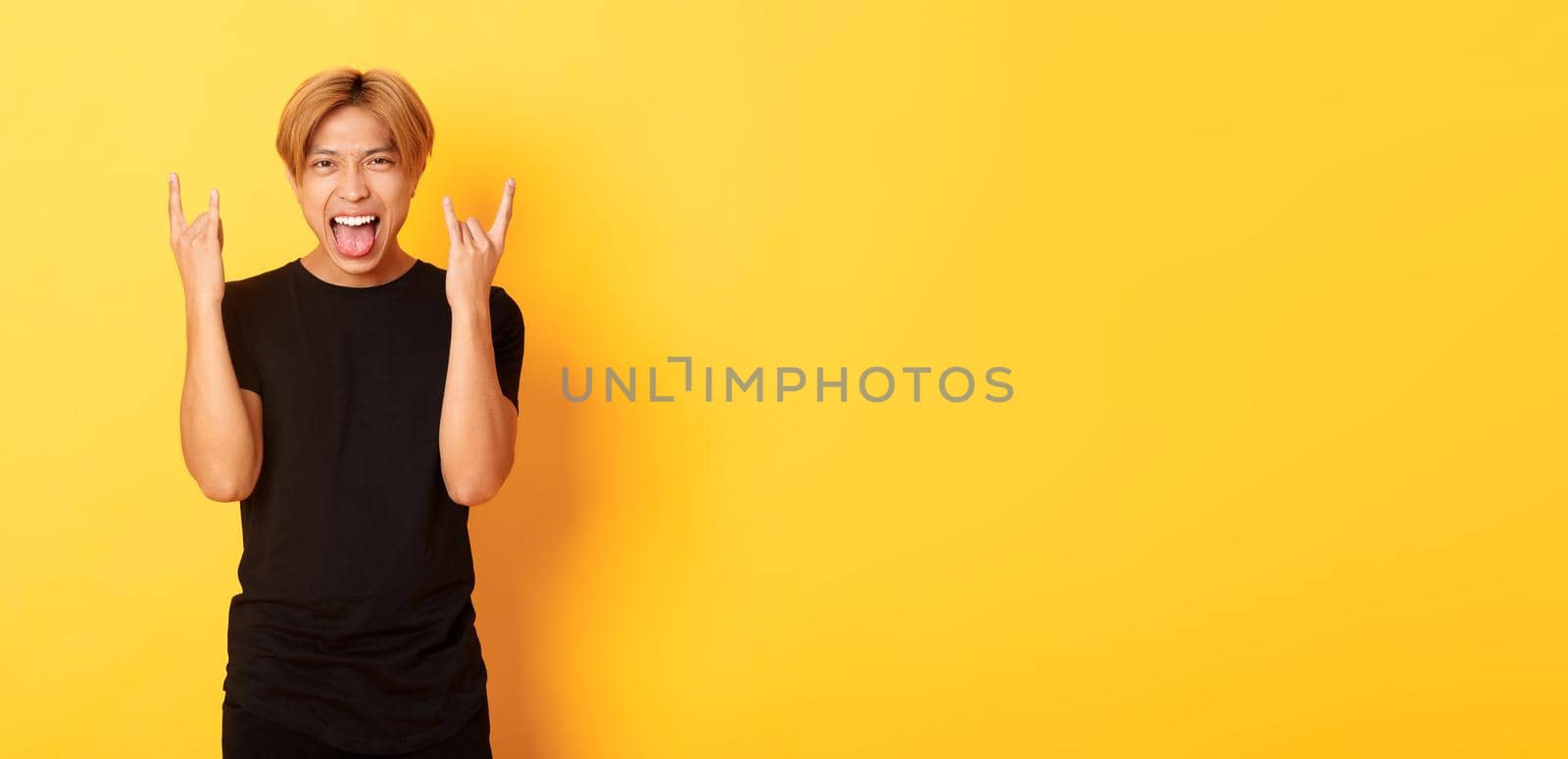Portrait of joyful handsome asian blond guy having fun, showing rock-n-roll gesture and stick tongue carefree, standing yellow background.