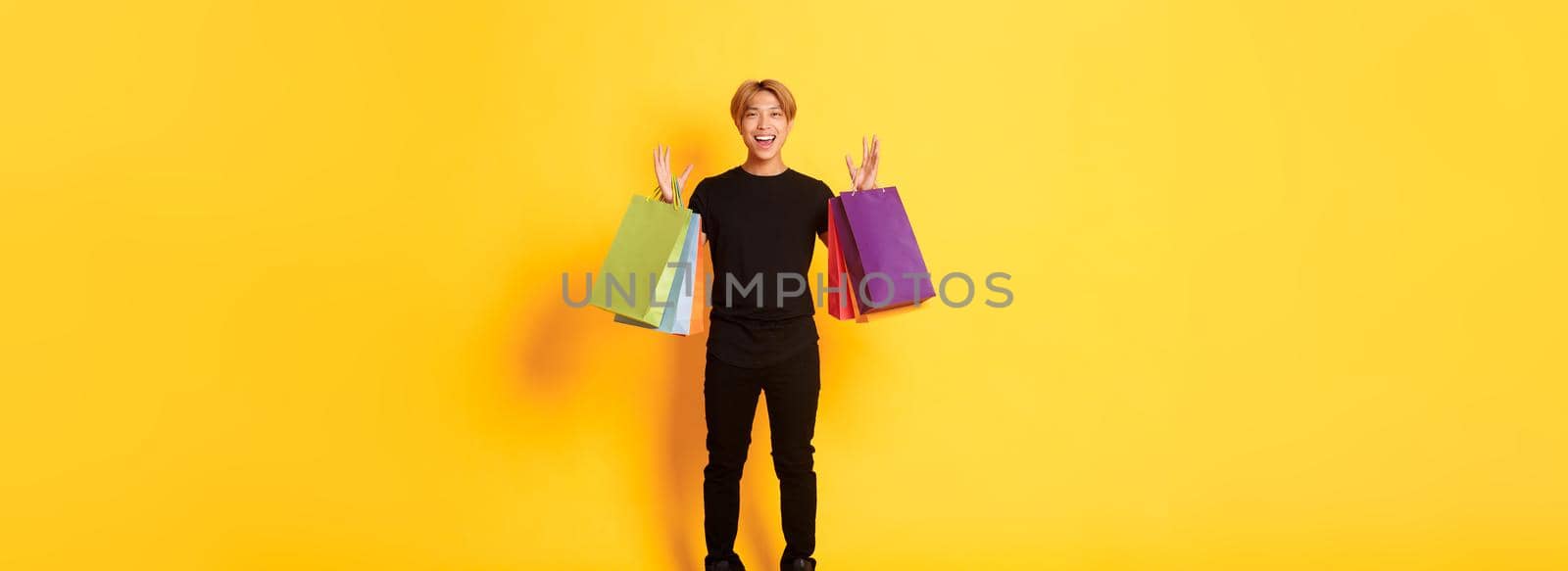 Full-length of happy handsome asian guy on shopping, holding bags and smiling, yellow background.