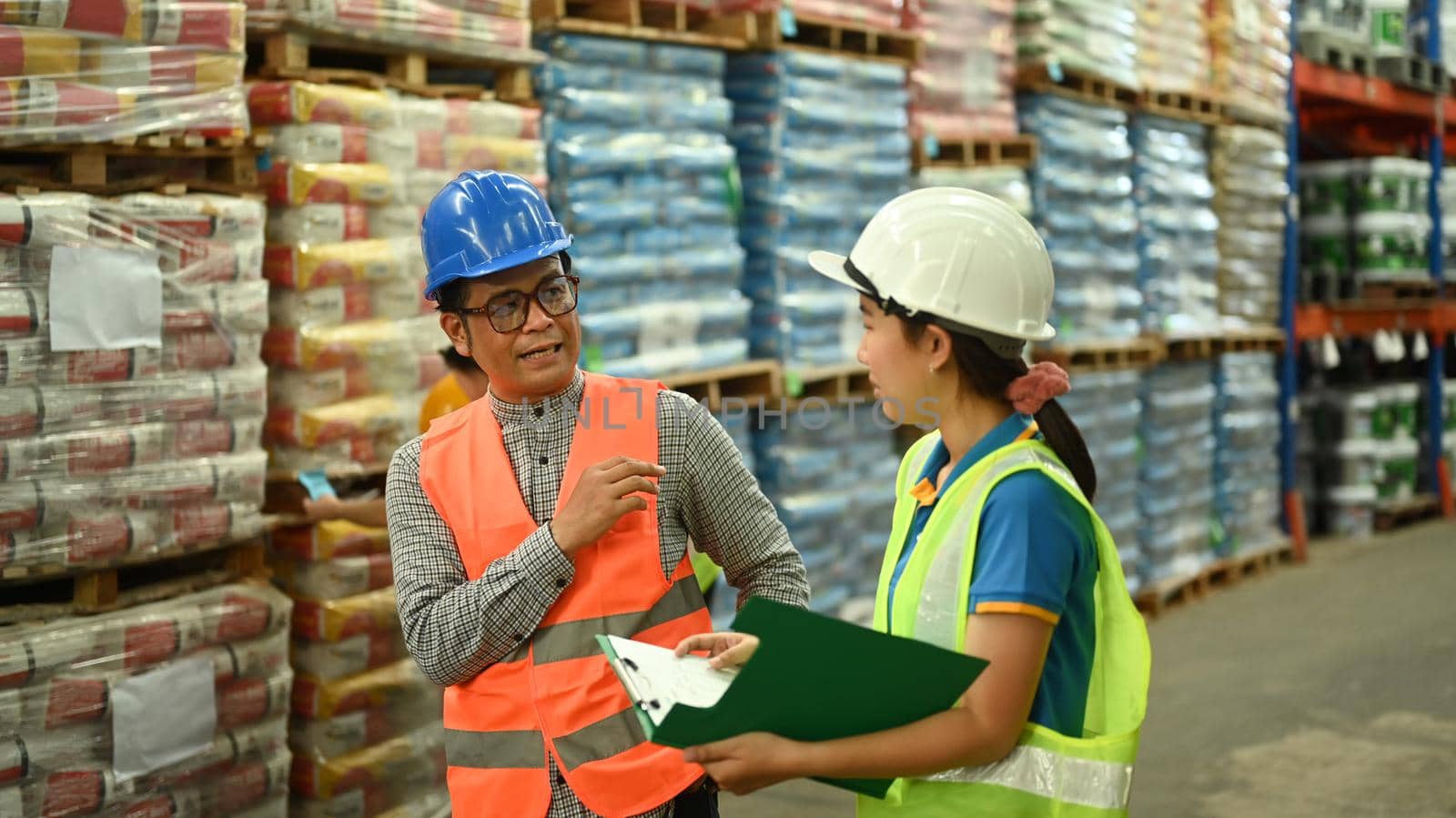 Senior male manager and woman warehouse worker discussing work while walking in aisle between rows of tall shelves full of packed boxes by prathanchorruangsak