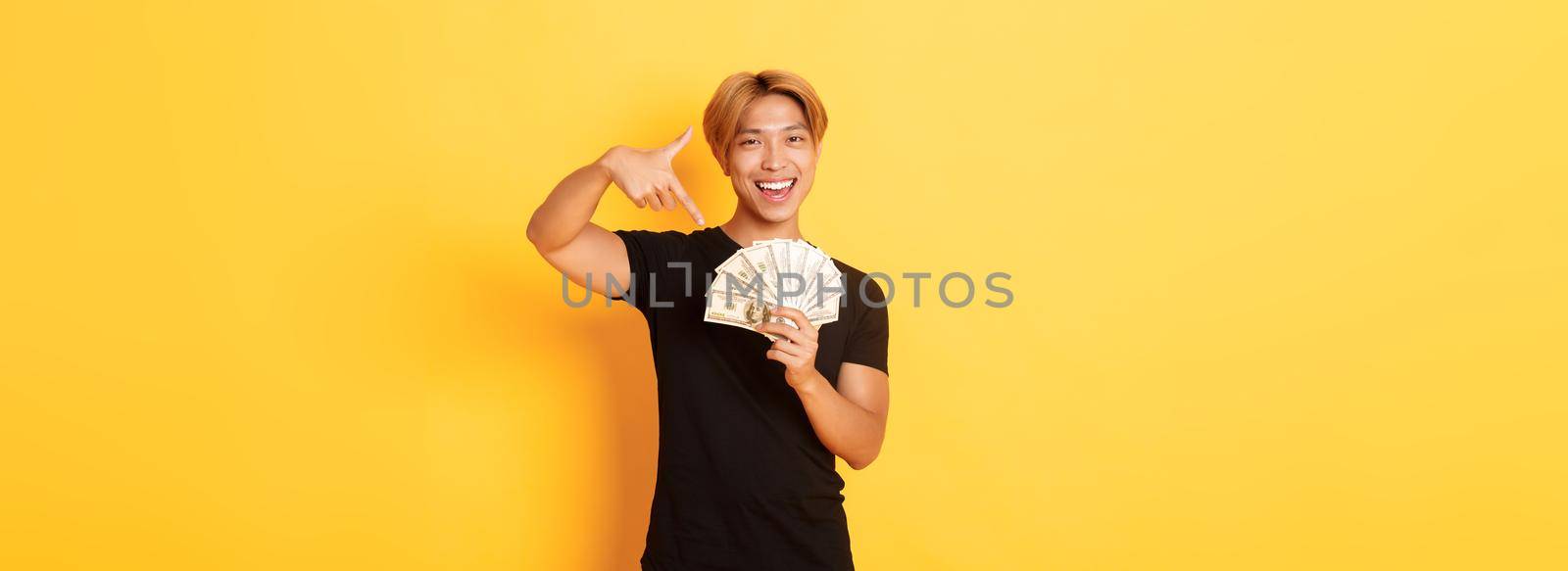 Sassy good-looking korean blond guy, smiling happy and pointing finger at money, winning cash, standing yellow background.