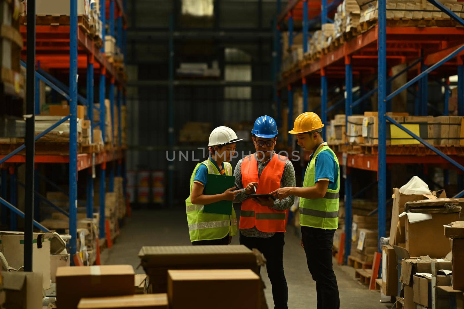 Warehouse workers and managers analyzing newly arrived goods on digital tablet while standing between rows of tall shelves.