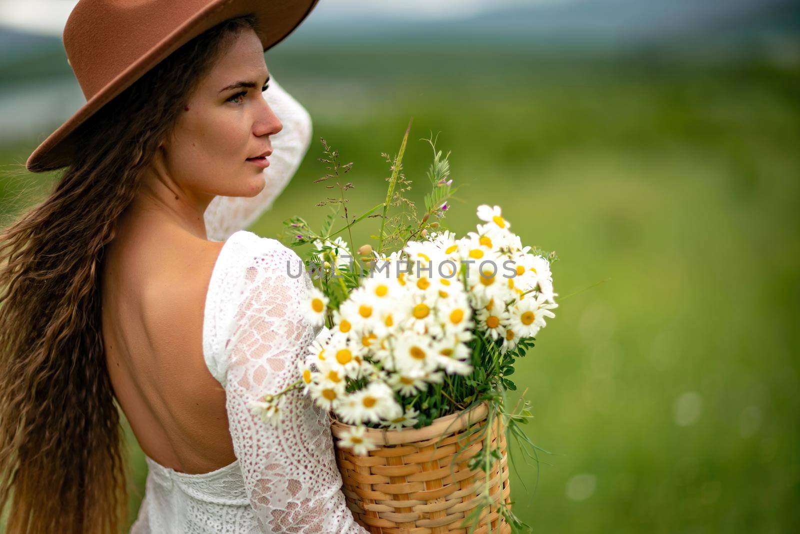 A middle-aged woman in a white dress and brown hat stands on a green field and holds a basket in her hands with a large bouquet of daisies. In the background there are mountains and a lake