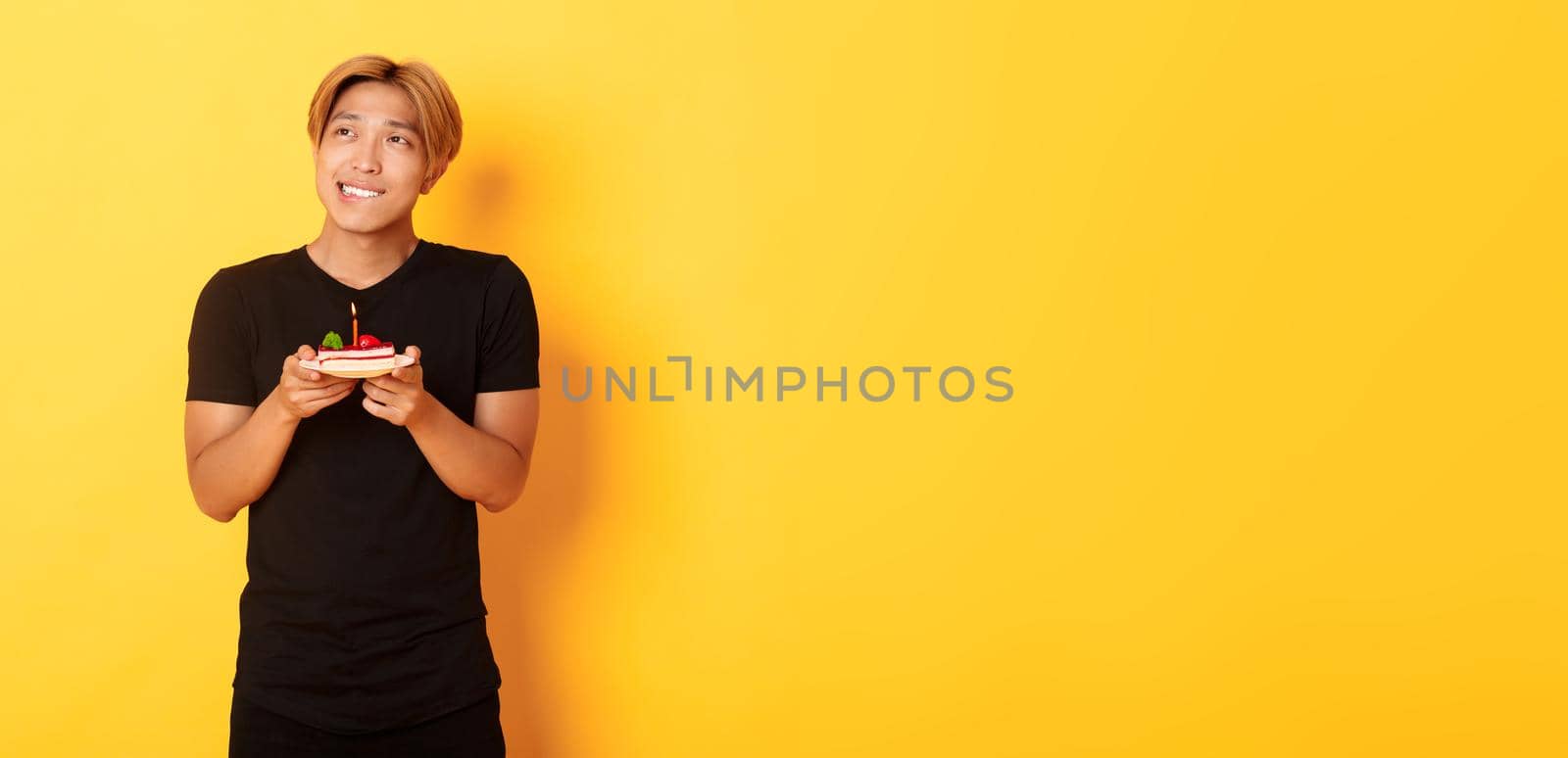 Portrait of handsome dreamy asian guy looking upper left corner and thinking, making wish while celebrating birthday and holding b-day cake, yellow background by Benzoix