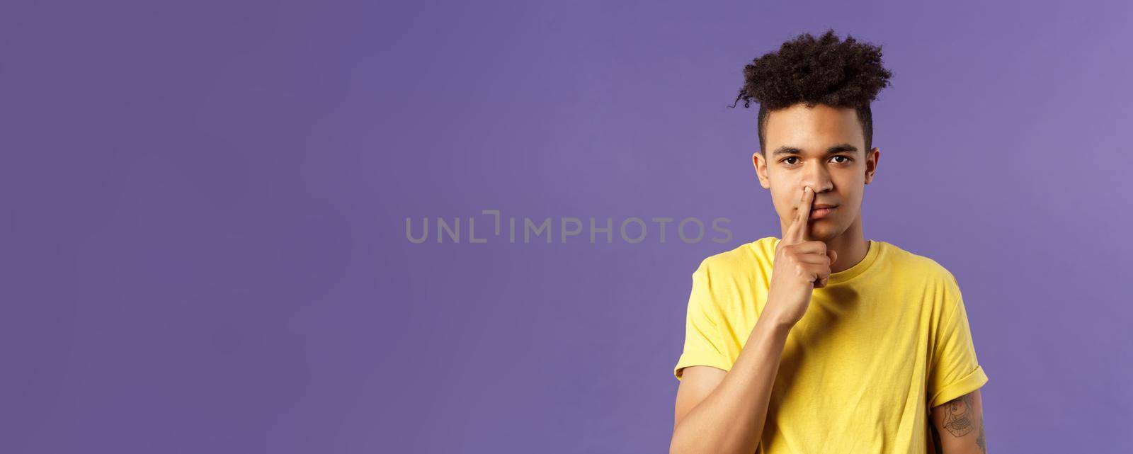 Close-up portrait of funny and unappropriate young hispanic teenage guy picking nose with finger, smiling and looking at camera, standing purple background, have bad habit.