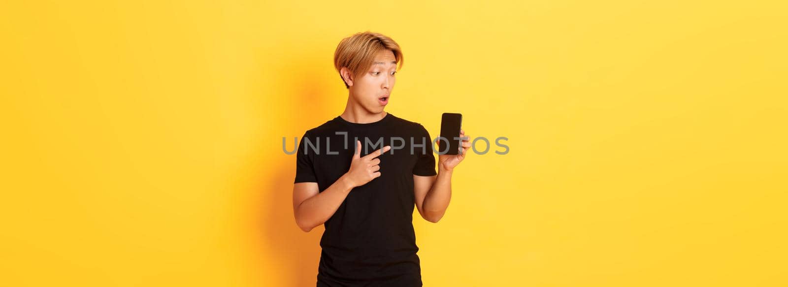 Portrait of handsome asian man showing something on smartphone screen, pointing at mobile phone display, standing over yellow background by Benzoix