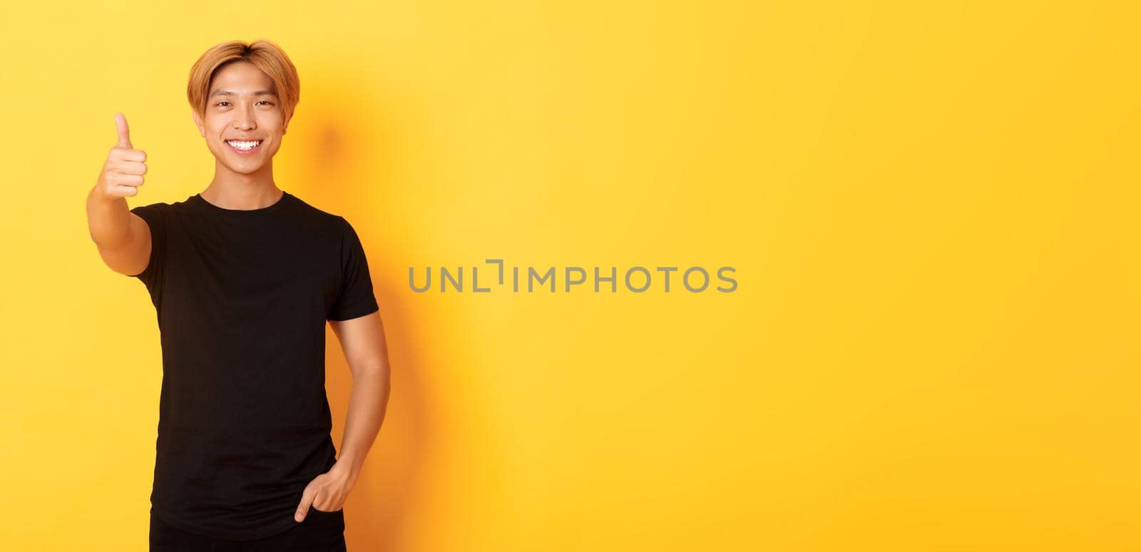 Portrait of satisfied asian man with blond hair, standing over yellow background and showing thumbs-up.