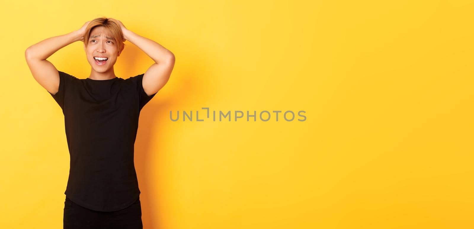 Portrait of young asian guy panicking, looking anxious and upset, holding hands on head and grimacing, standing over yellow background by Benzoix