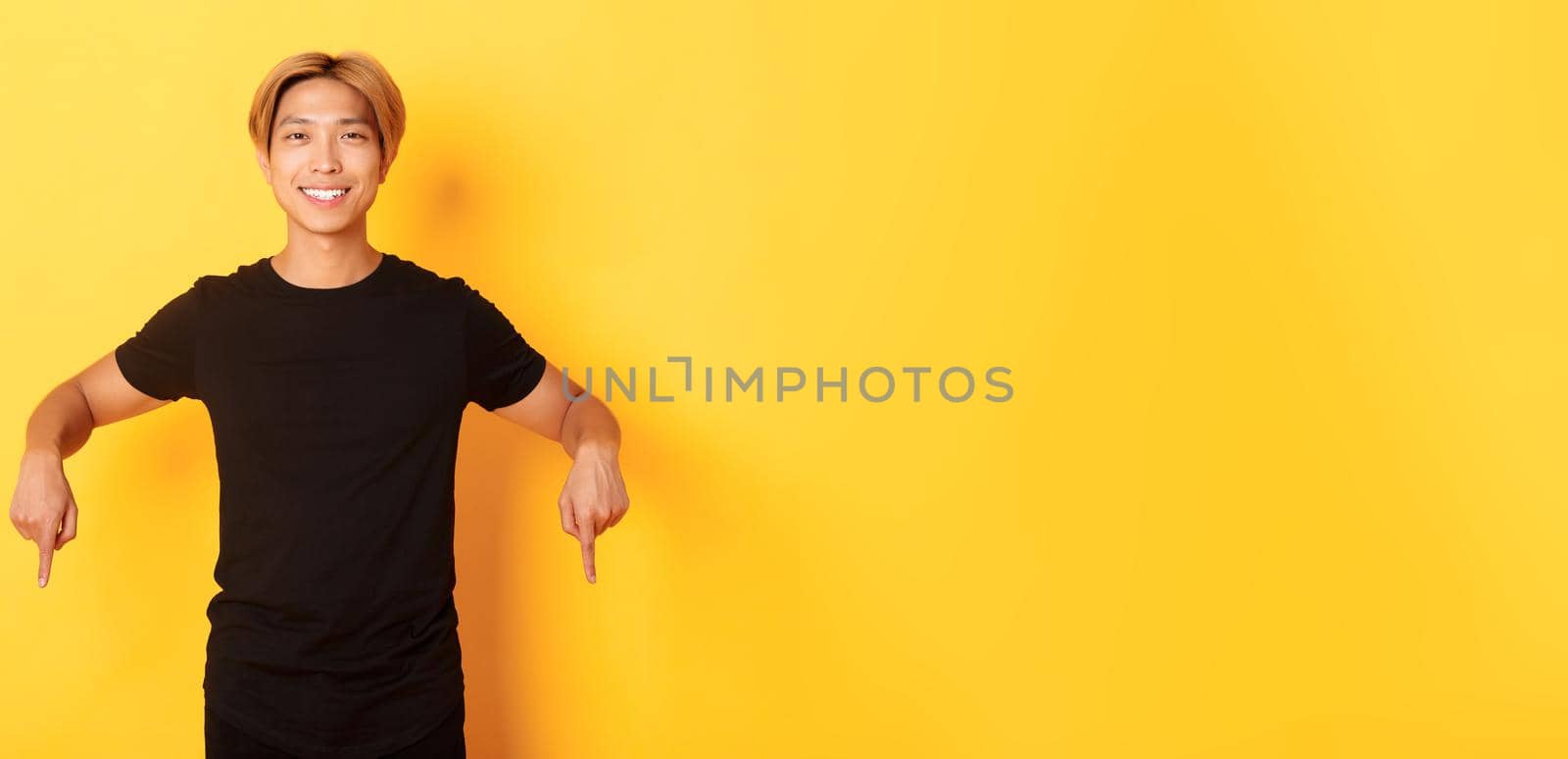 Portrait of smiling attractive korean guy with blond hair, wearing black outfit, pointing fingers down, showing way, standing yellow background by Benzoix