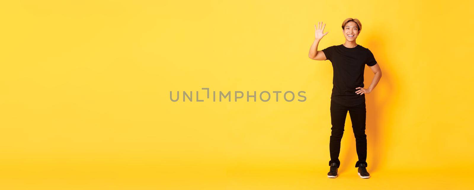 Full-length of friendly handsome asian man waving hand in hello, smiling and saying hi over yellow background.