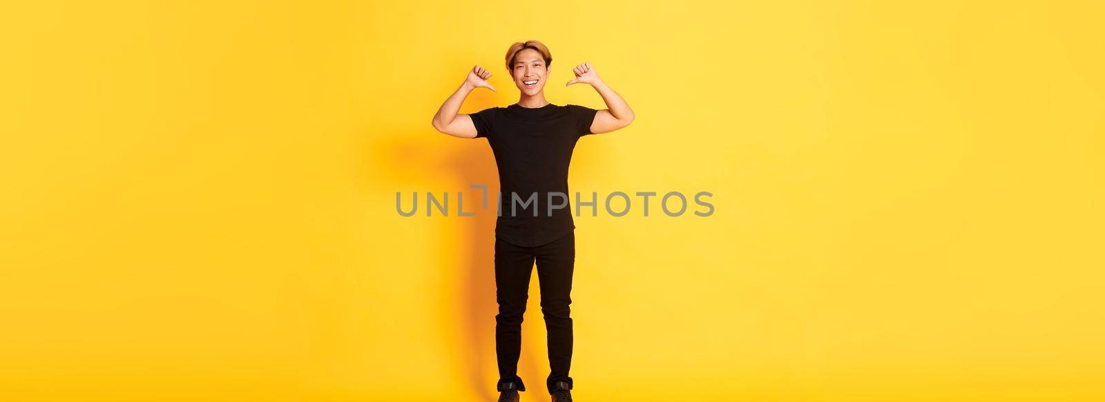 Confident attractive blond asian guy, smiling sassy and pointing at himself, standing over yellow background by Benzoix