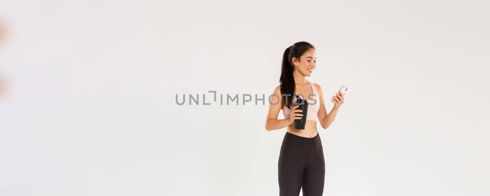 Full length of smiling fit and healthy female fitness coach, asian sportswoman checking water tracker reminder in smartphone, using application during workout in gym, white background.