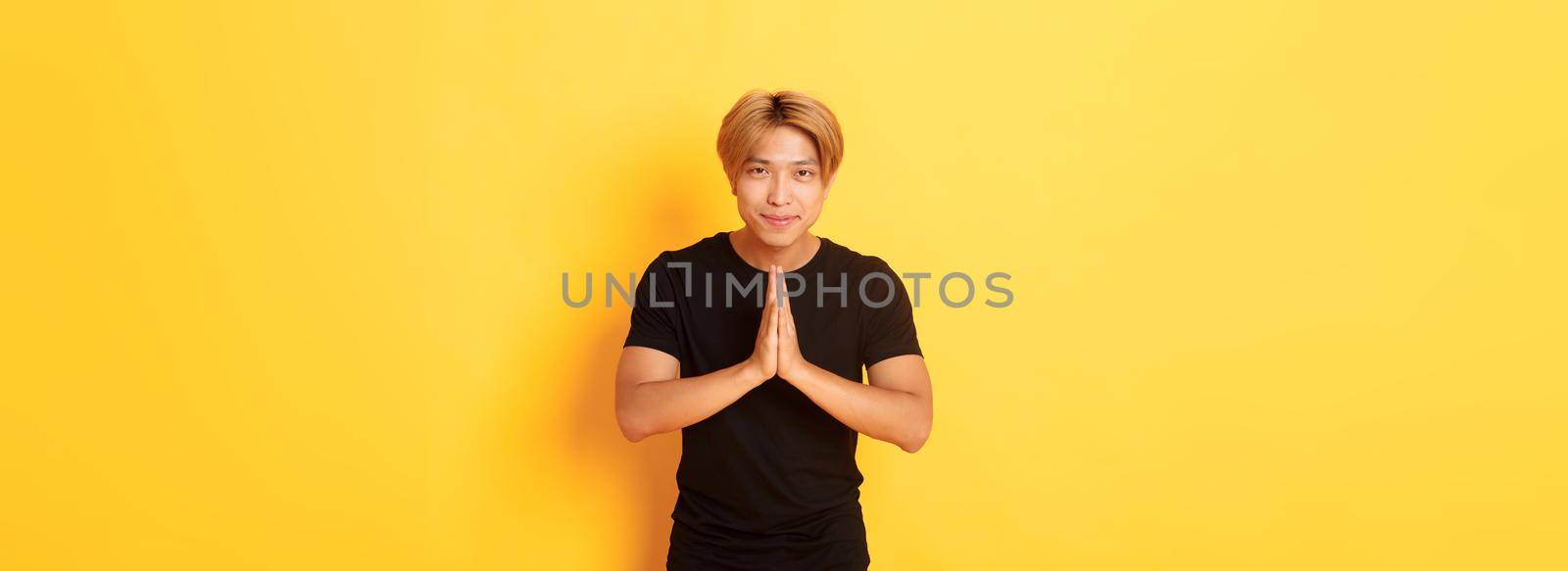 Portrait of friendly and polite asian blond guy, clasp hands together in namaste gesture, bowing and greeting person, standing over yellow background by Benzoix