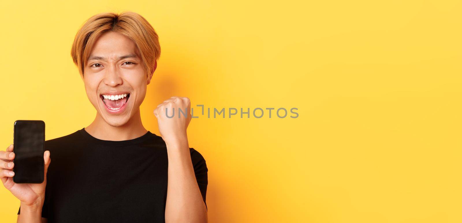 Close-up of happy rejoicing asian guy showing smartphone screen and saying yes, fist pump as triumphing, winning or achieve goal, yellow background by Benzoix