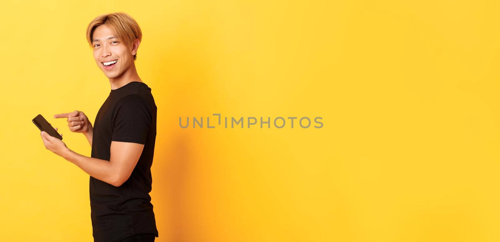 Portrait of pleased smiling handsome asian man, standing in profile and pointing finger at smartphone, recommend app, standing yellow background.