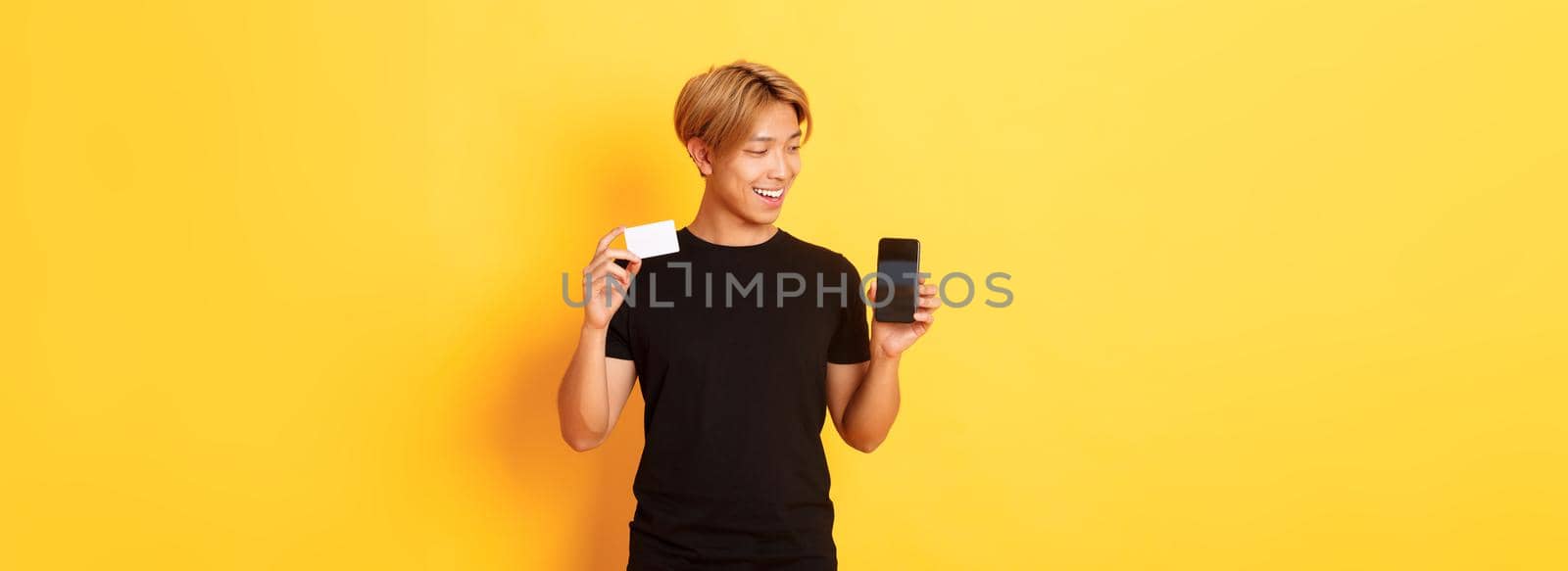 Portrait of pleased handsome asian guy, showing smartphone screen and credit card, looking at mobile phone display, yellow background.