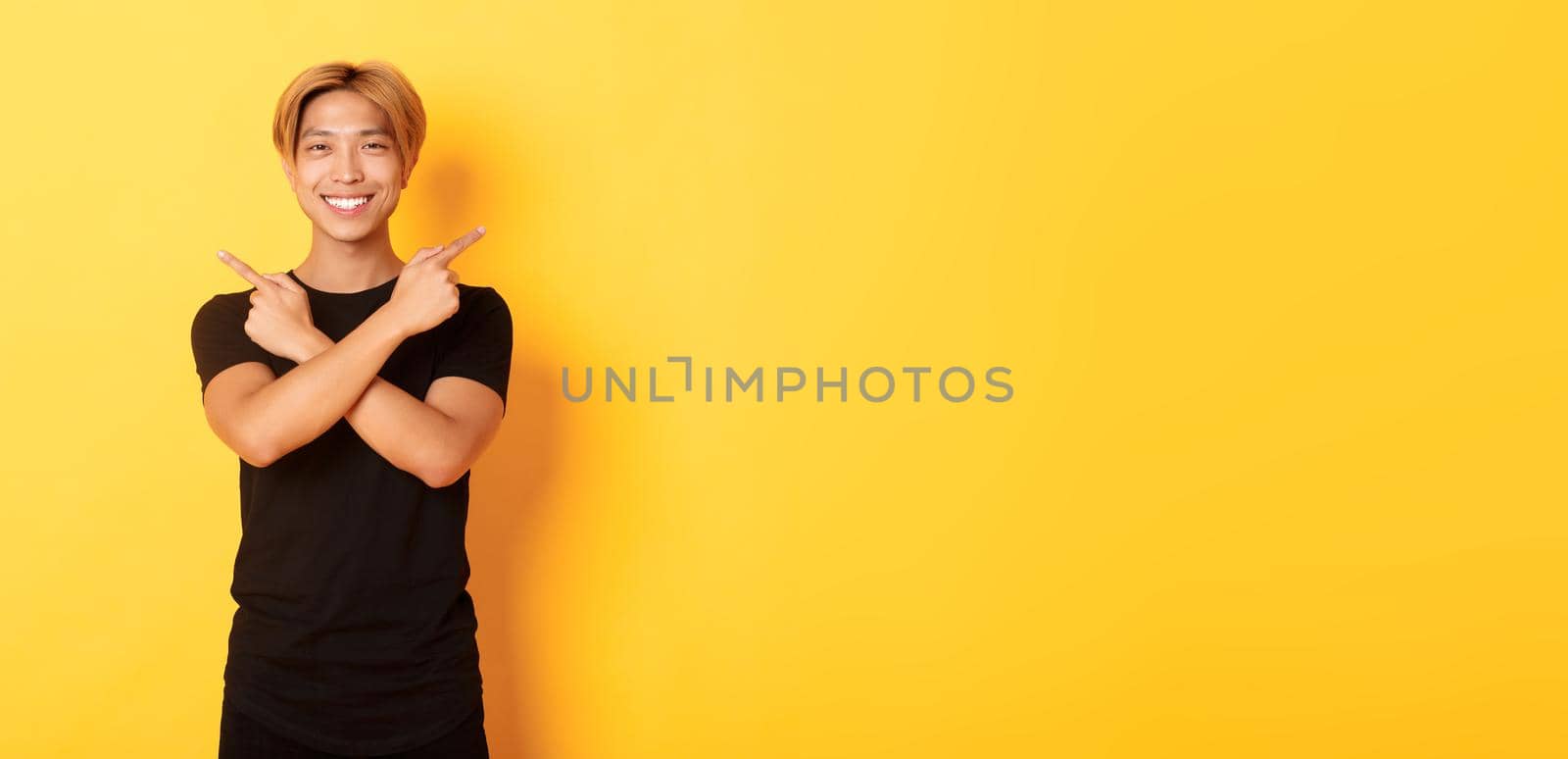 Portrait of handsome determined blond asian guy, student pointing fingers sideways, showing two variants, standing yellow background.