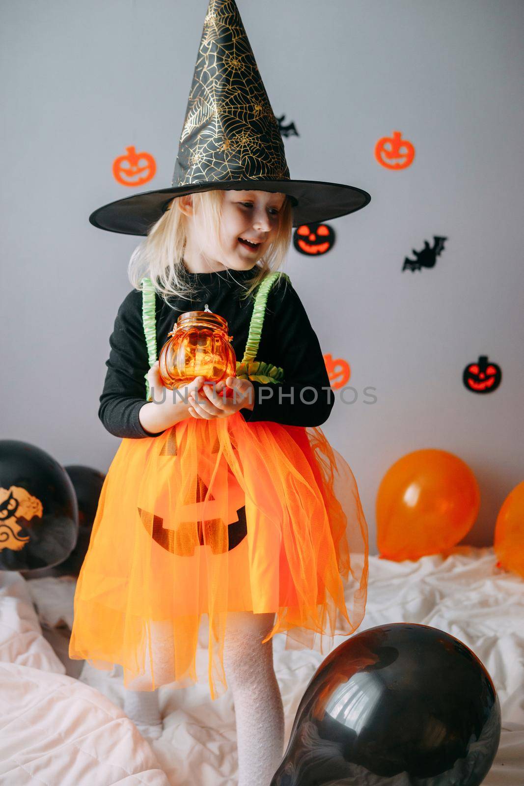 Children's Halloween - a girl in a witch hat and a carnival costume with airy orange and black balloons at home. Ready to celebrate Halloween.