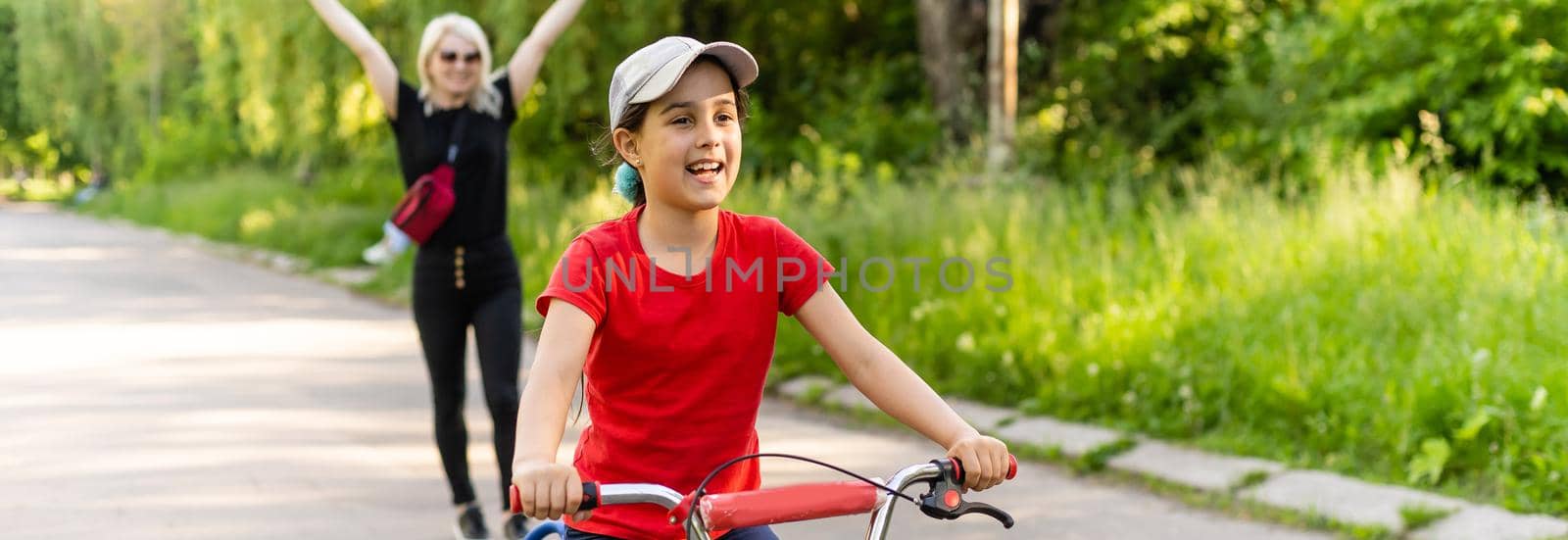 Beautiful young mother teaching her daughter to ride a bicycle. Both smiling, summer park in background, active family concept.