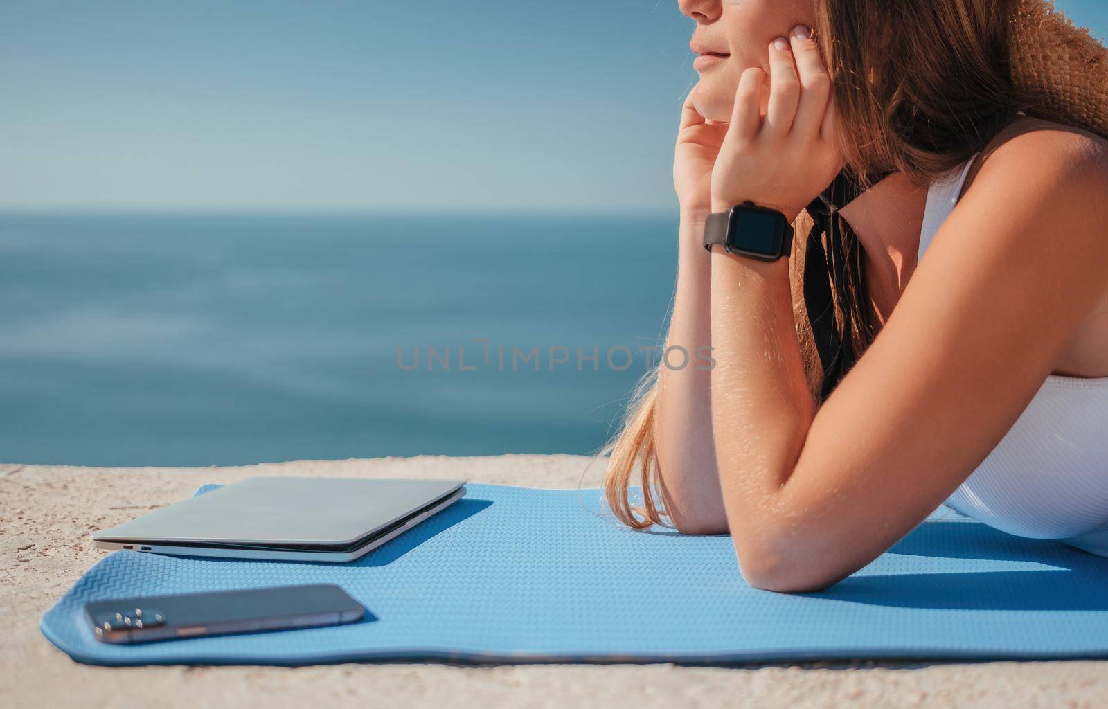 Successful business woman in yellow hat working on laptop by the sea. Pretty lady typing on computer at summer day outdoors. Freelance, travel and holidays concept.
