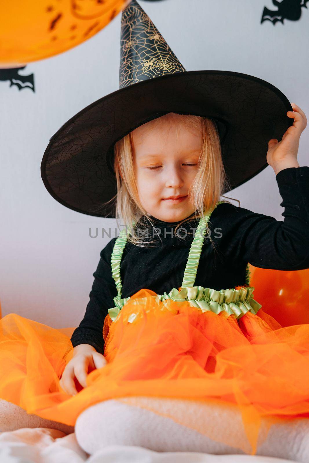 Children's Halloween - a girl in a witch hat and a carnival costume with airy orange and black balloons at home. Ready to celebrate Halloween.