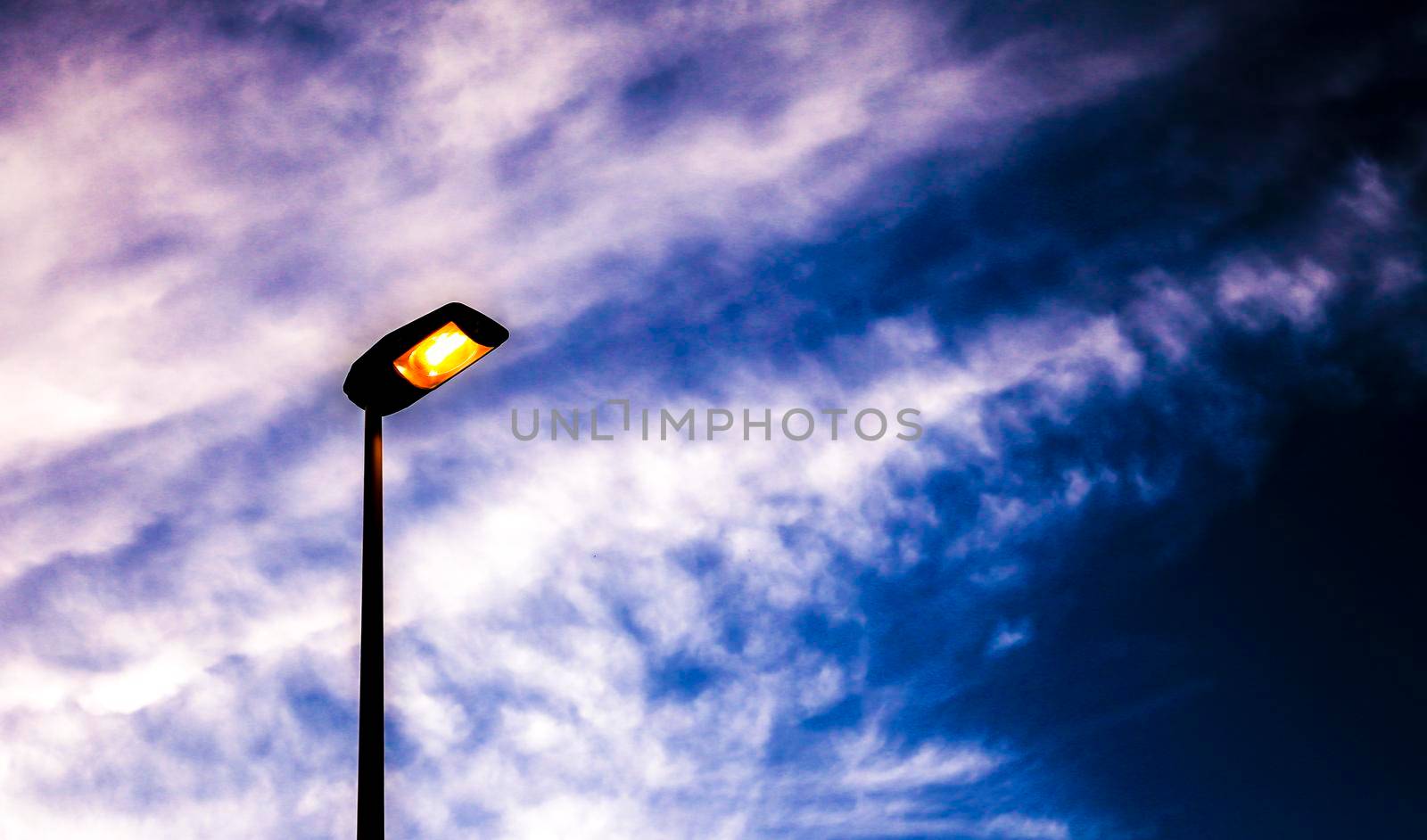 Lamppost illuminating with orange light the afternoon. Blue sky and white clouds in the background.