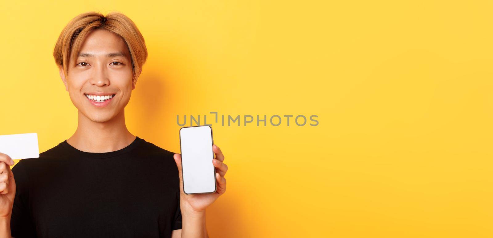 Close-up of smiling handsome asian guy showing smartphone screen and credit card, standing yellow background.