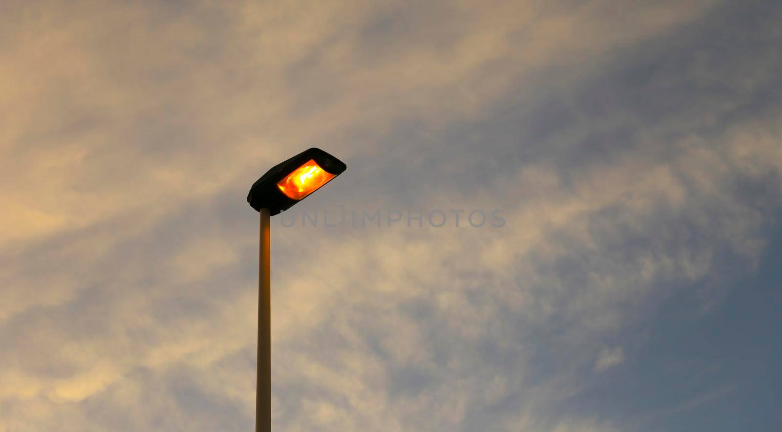 Lamppost illuminating with orange light the afternoon. Blue sky and white clouds in the background.