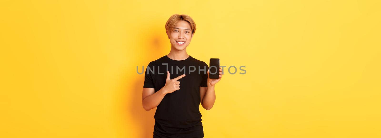 Portrait of handsome asian man showing something on smartphone screen, pointing at mobile phone display, standing over yellow background.