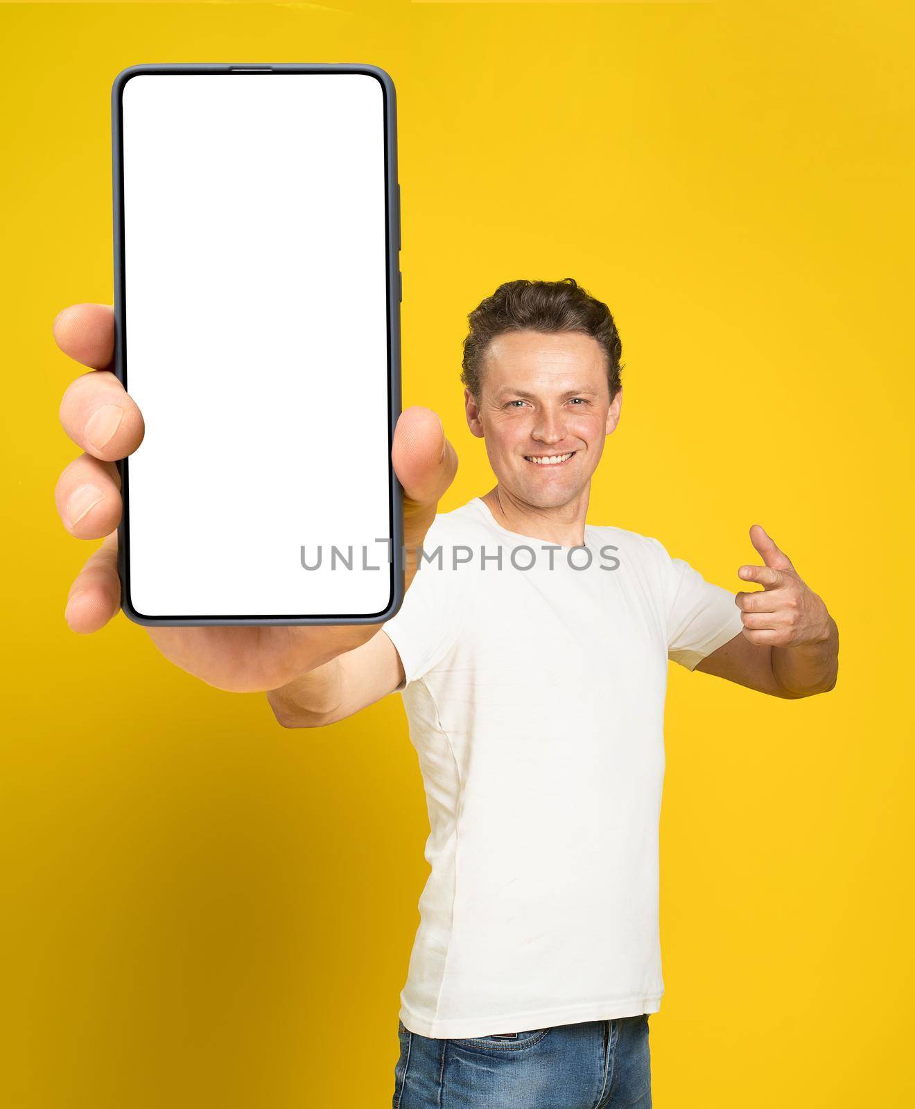 Holding huge smartphone with white screen handsome man pointing at white empty screen, wearing white t-shirt and jeans isolated on yellow background. Mobile app advertisement, great offer.