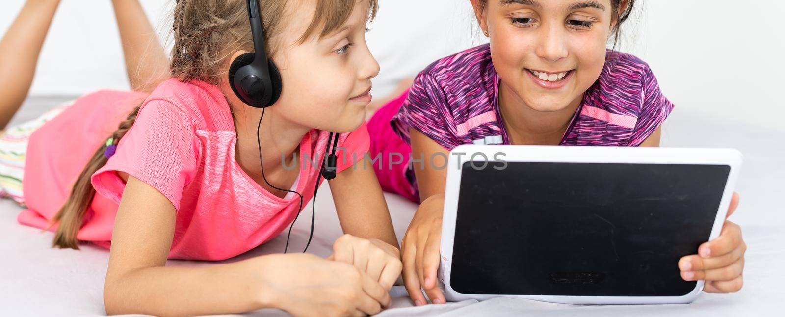 Two cute little girls are using a digital tablet and smiling while lying on bed in children's room.