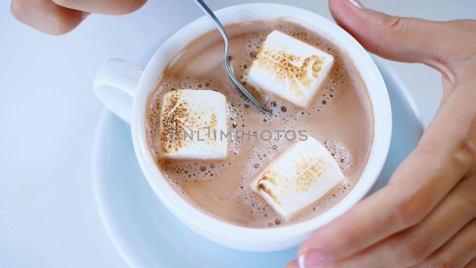 close-up, top view, white cup with cappuccino, with thick creamy foam and marshmallows. a woman's hand slowly stirs the drink with a teaspoon. High quality photo