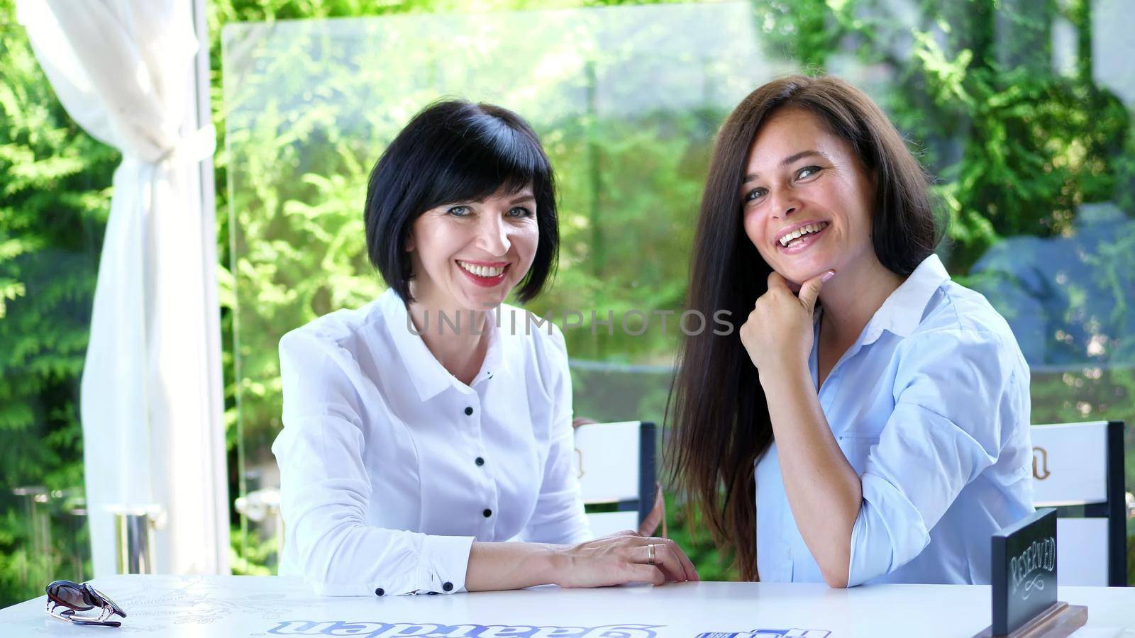 in the summer, on a terrace in a cafe, two forty-year-old beautiful brunettes of a woman are chatting cute, they are dressed in blond blouses. High quality photo