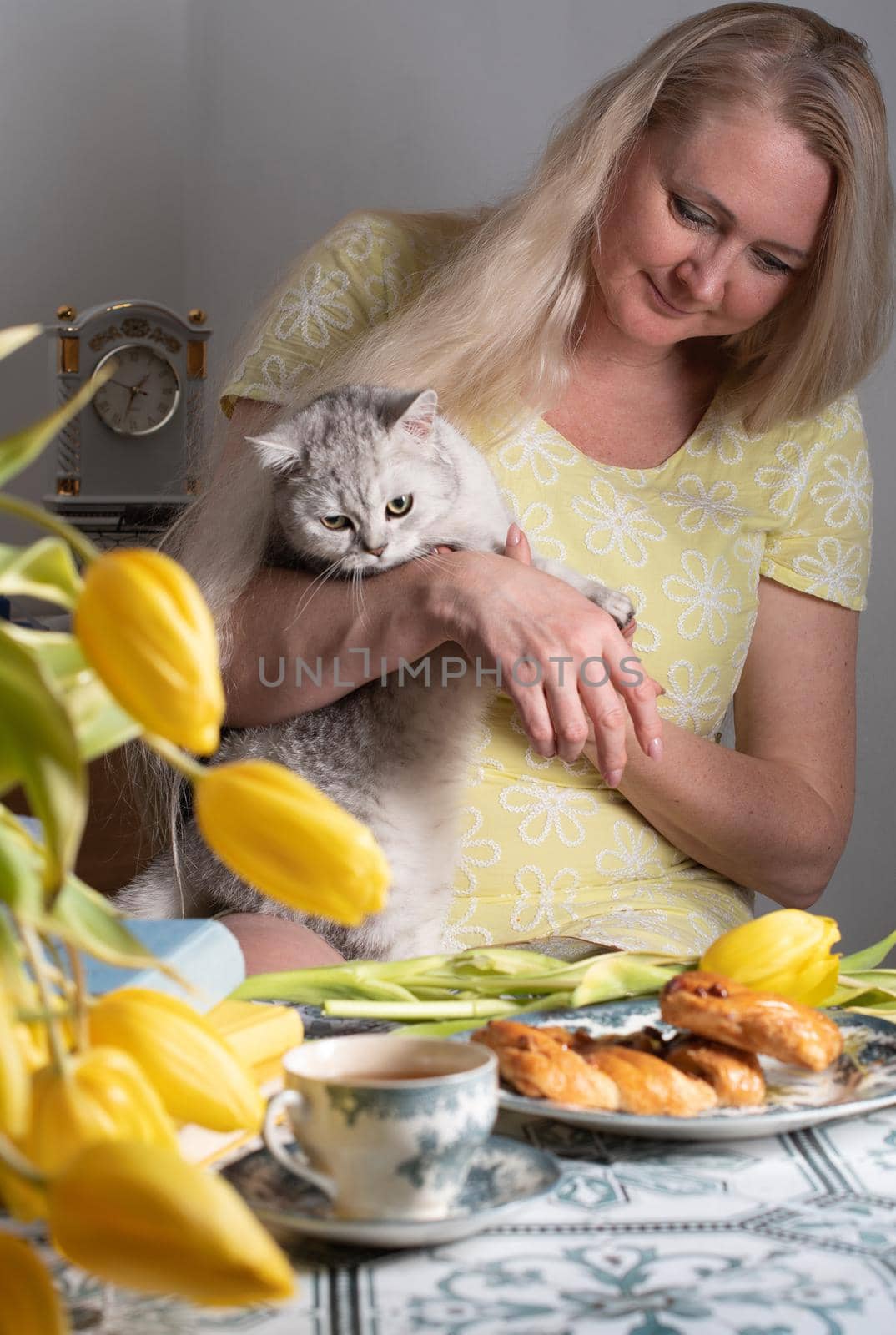 blonde middle-aged hugging a kitten and drinking tea, still life with yellow tulips and cakes a stack of books on the table. High quality photo