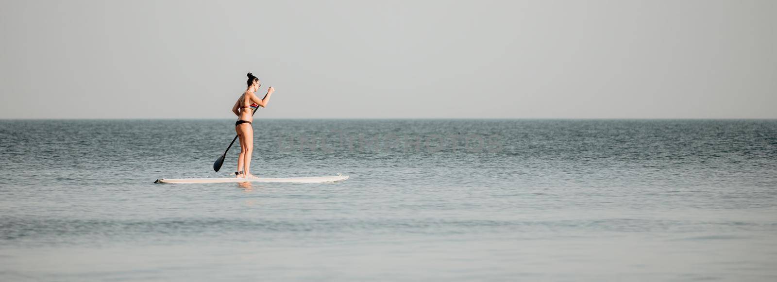 Sea woman sup. Silhouette of happy middle aged woman in rainbow bikini, surfing on SUP board, confident paddling through water surface. Idyllic sunset. Active lifestyle at sea or river. by panophotograph