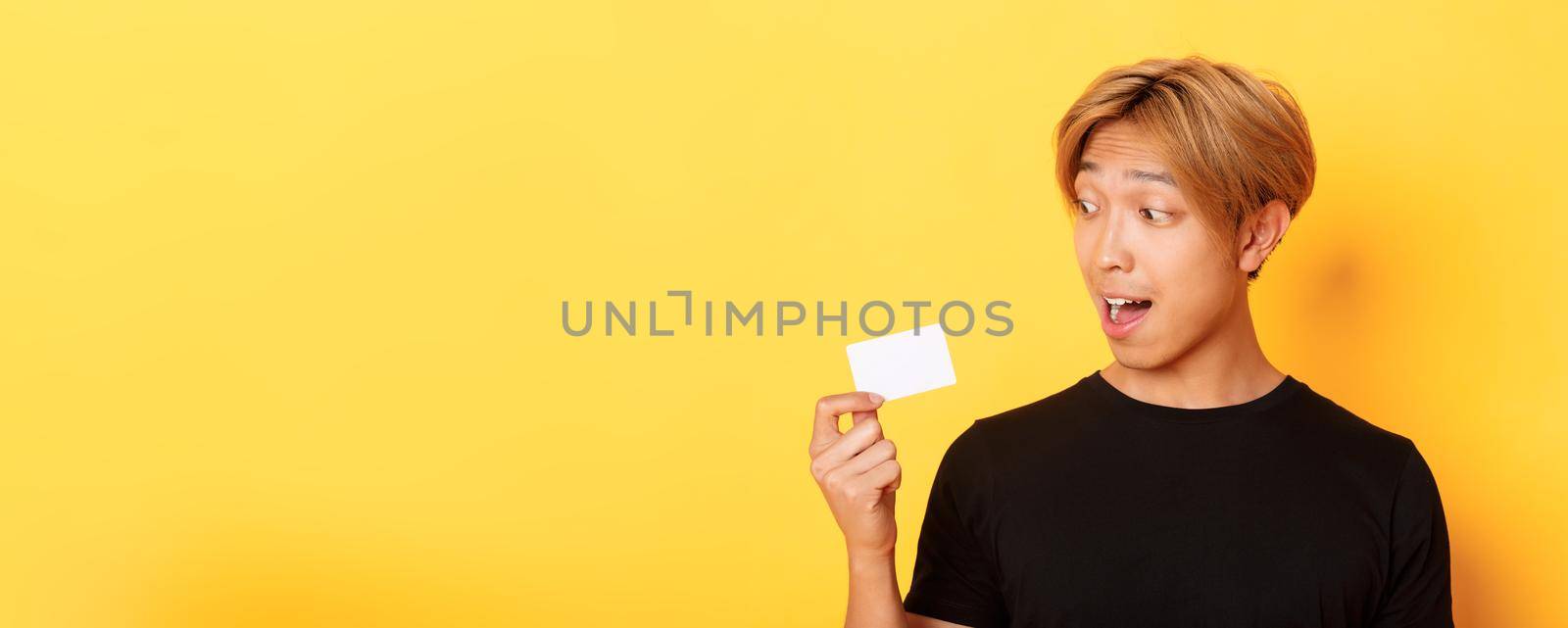 Close-up of amazed asian guy talking about credit card, standing over yellow background.