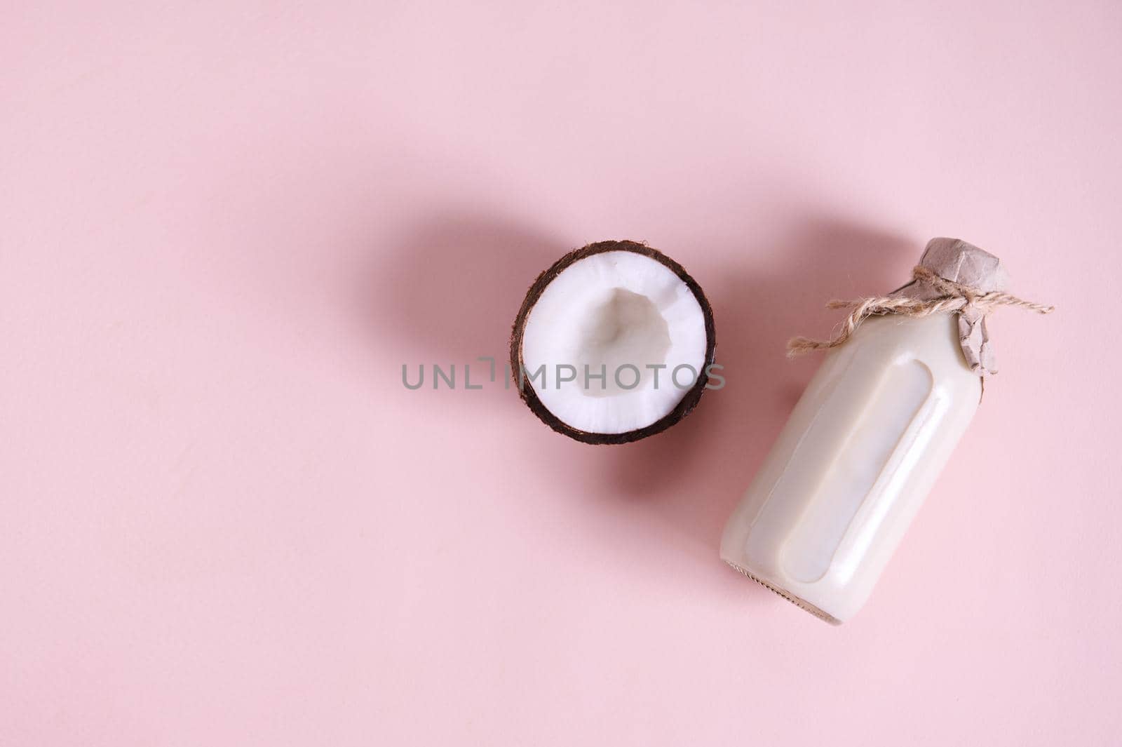 Flat lay composition with a half of raw coconut, lying next to a glass bottle with organic vegan plant based milk, isolated on pink background with copy space for advertising text. Still life