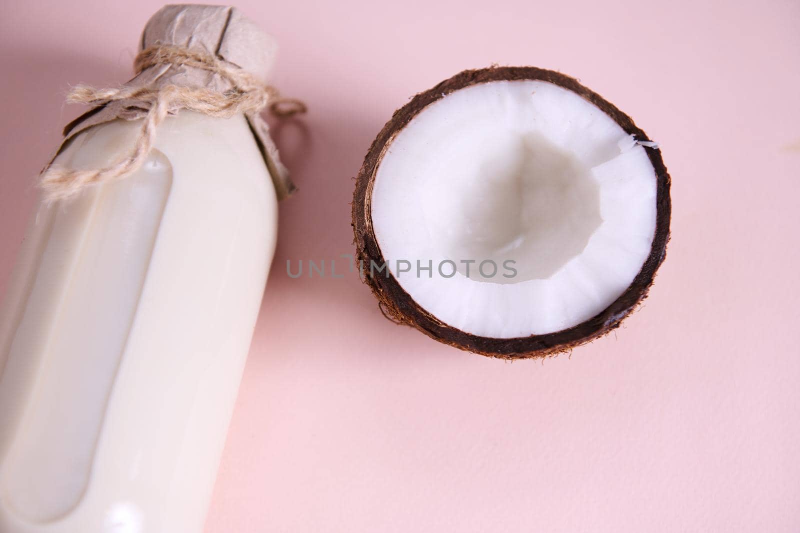 Close-up of a glass bottle of organic raw vegan milk with a craft paper tying with a bow on a lid, and a half of coconut, isolated over pink background with copy ad space. Healthy food and drink