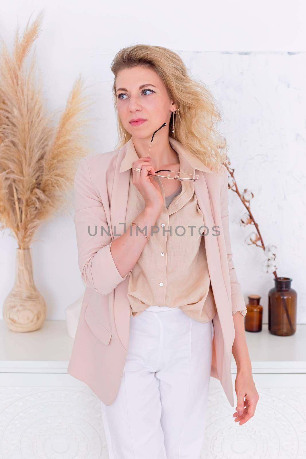 A beautiful woman with blond hair, stands in a light pink jacket in the interior of a white room, look to the side . Copy space. Vertical