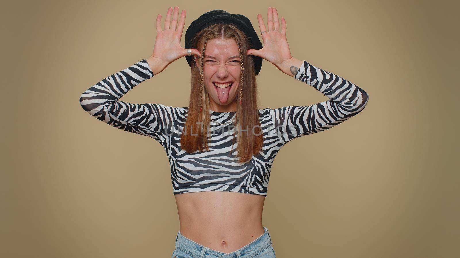 Lovely pretty funny woman in crop top making playful silly facial expressions and grimacing, fooling around, showing tongue. Adult stylish female girl isolated alone on beige studio background indoors