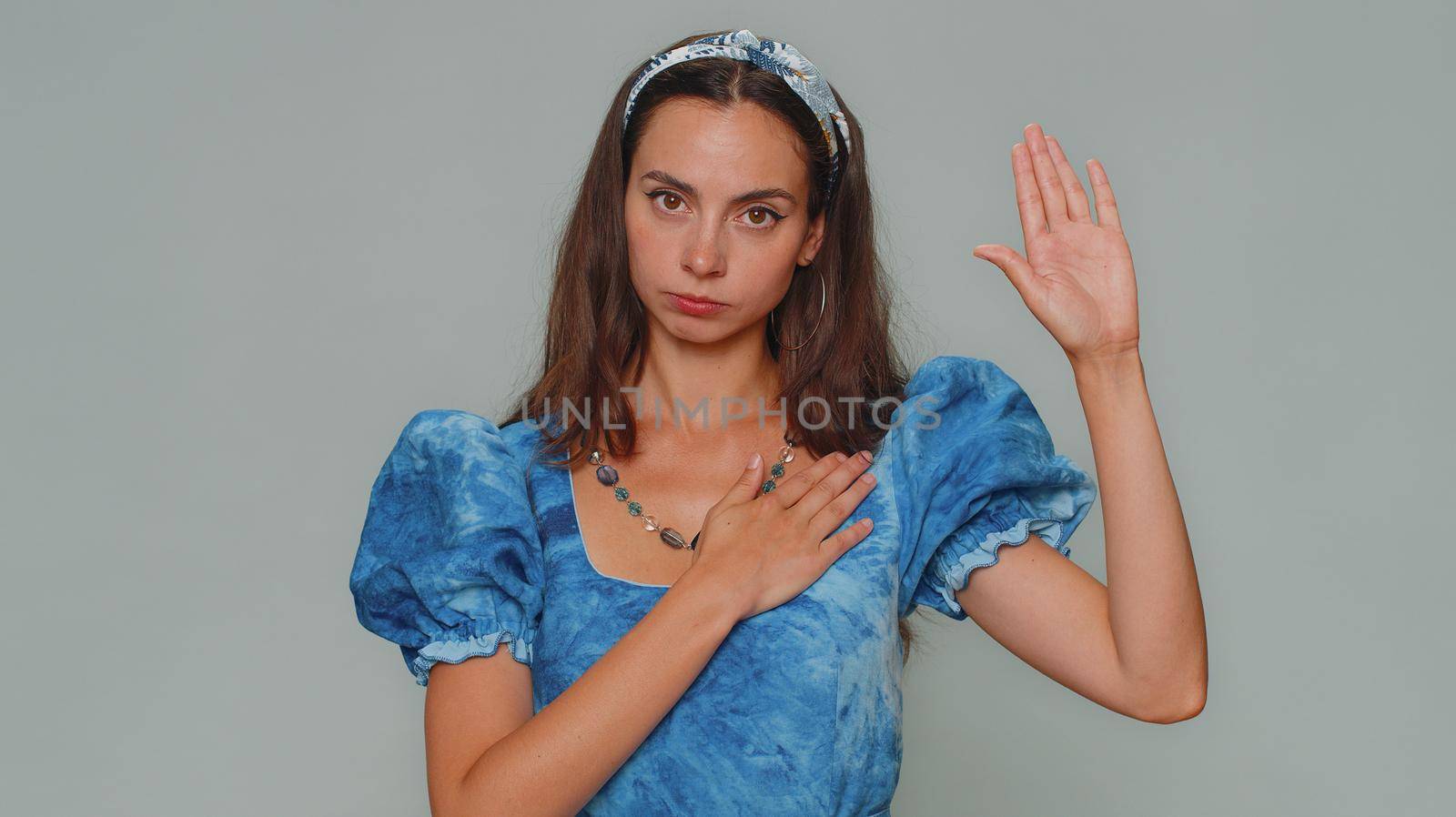 I swear to be honest. Sincere responsible pretty young woman raising hand to take oath, promising to be honest and to tell truth, keeping hand on chest. Adult stylish girl isolated on gray background