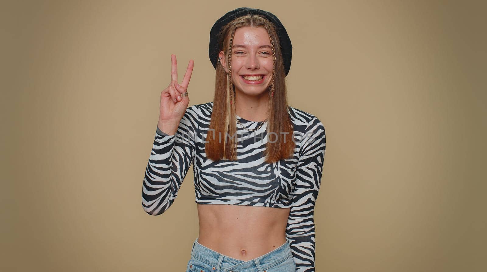 Lovely happy young woman showing victory sign, hoping for success and win, doing peace gesture, smiling with kind optimistic expression. Adult stylish girl isolated on beige studio background indoors