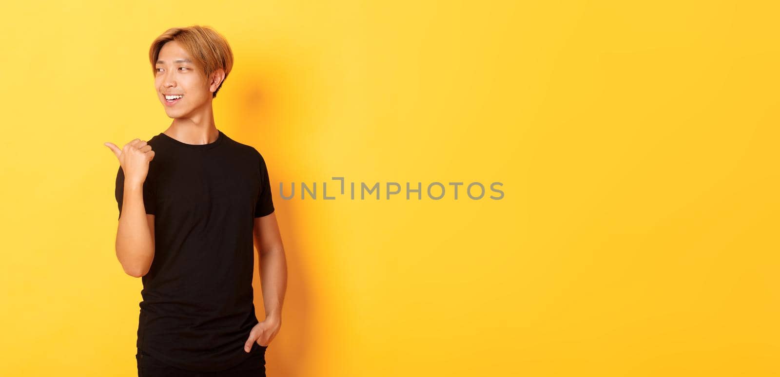 Portrait of pleased thoughtful asian guy, looking and pointing finger left at logo, standing yellow background by Benzoix