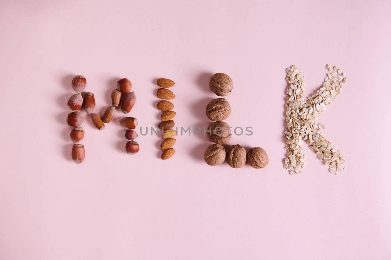 Flat lay. Lettering Milk using various healthy nuts, cereals on pink background with advertising copy space. Alternative organic raw vegan healthy drink made from oats, almonds, walnuts and hazelnuts
