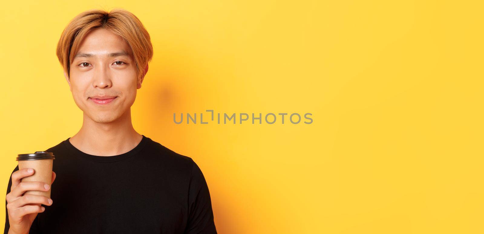 Close-up of handsome blond asian man drinking coffee and smiling pleased, standing over yellow background by Benzoix