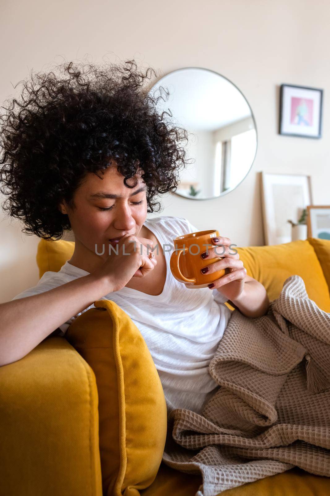 lll African american woman with sore throat coughing at home, sitting on the couch drinking healing herbal tea. Vertical by Hoverstock